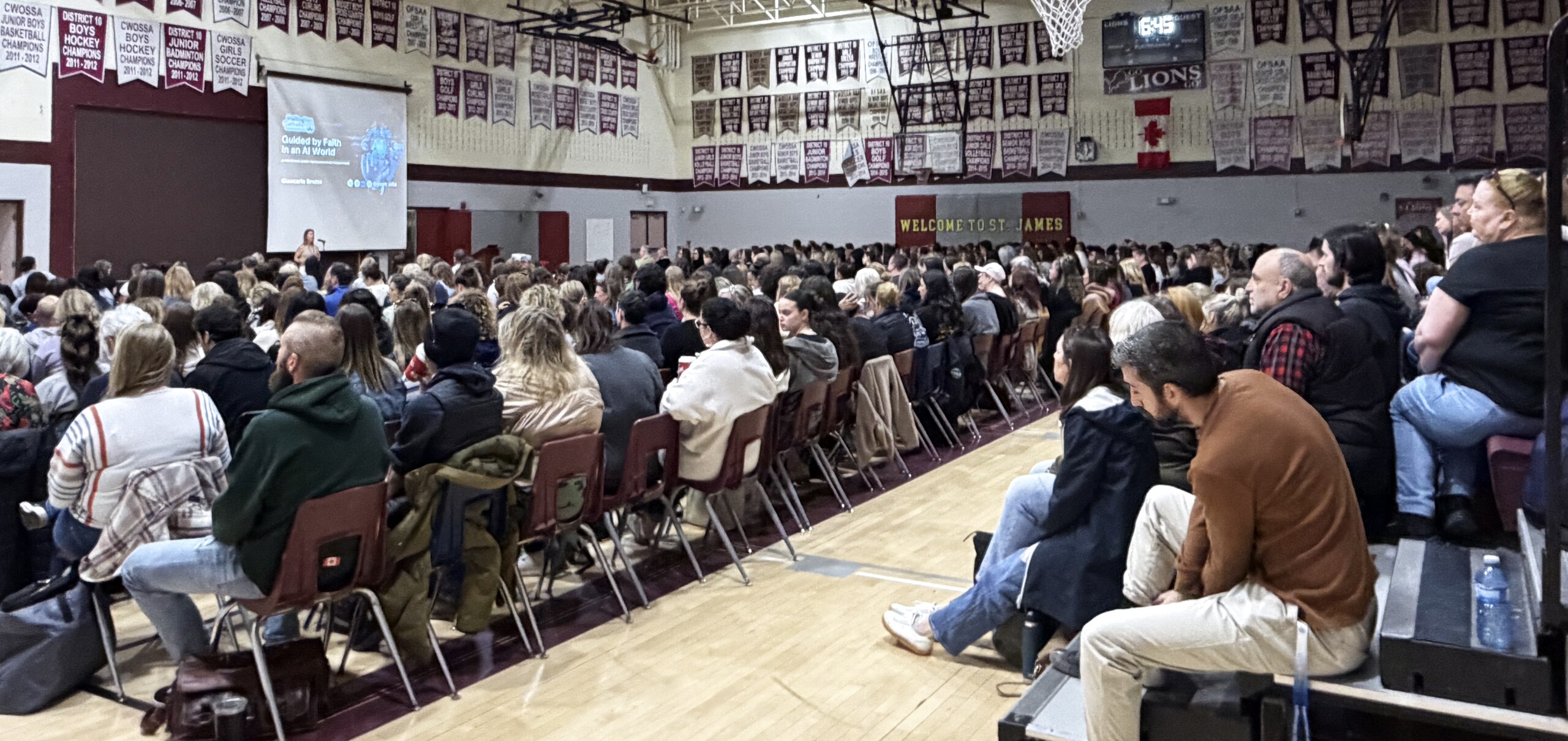 A large school community audience listens to a speaker in a gymnasium.